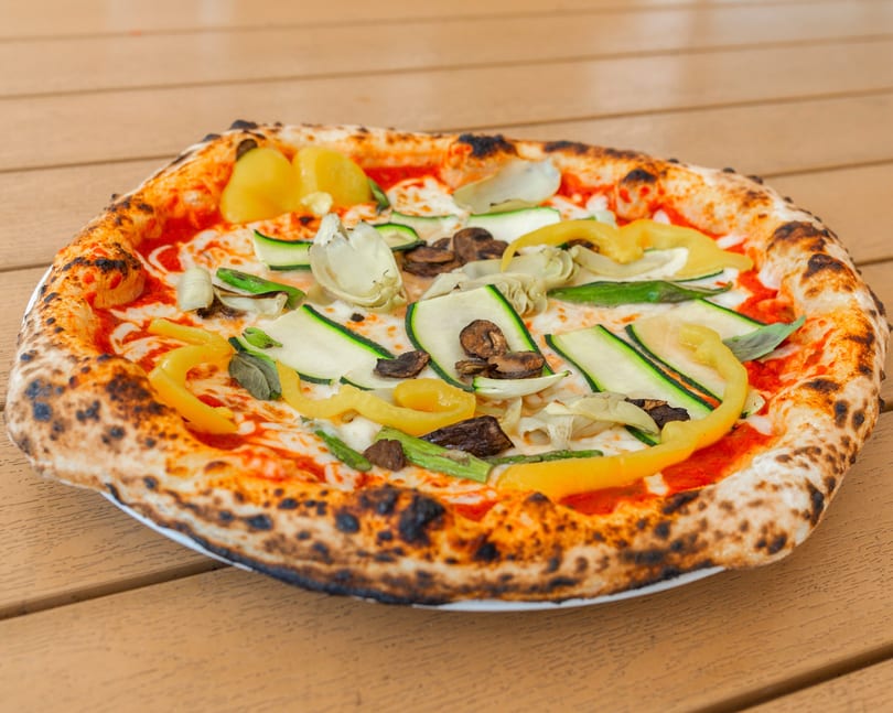Neapolitan pizza with zucchini, yellow peppers, mushrooms, and artichoke hearts on a wooden table.