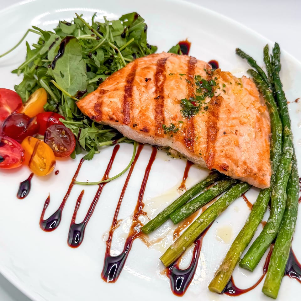 Grilled salmon with asparagus, arugula salad, cherry tomatoes, and balsamic glaze on a white plate.
