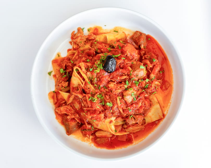 Pappardelle pasta with a rich red meat sauce, garnished with parsley and a black olive.
