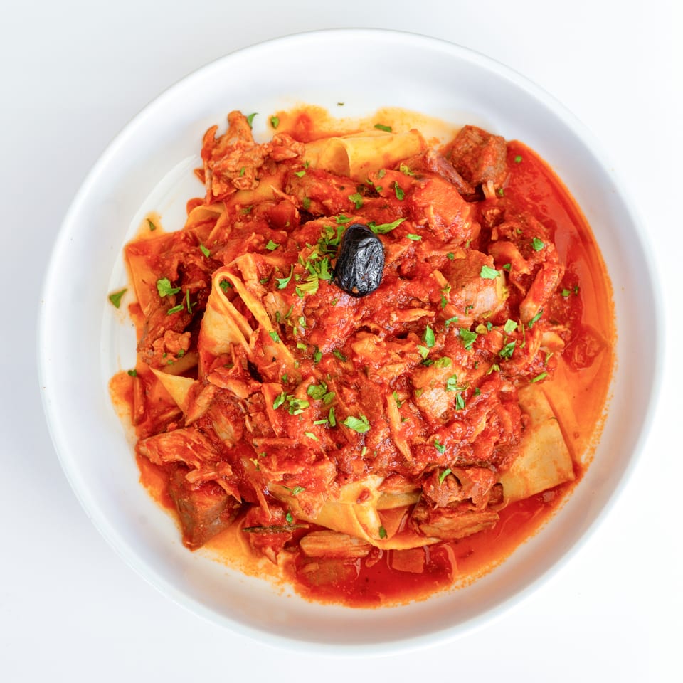 Pappardelle pasta with rich red tomato sauce, shredded meat, and a black olive in a white bowl.