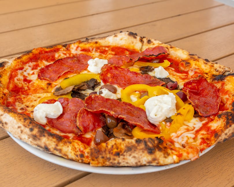Close-up of a pizza with pepperoni, yellow peppers, mushrooms, olives, and dollops of ricotta.