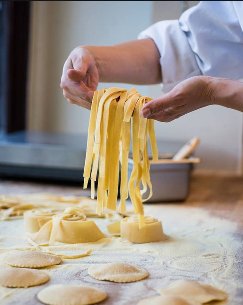 Chef's hands holding fresh tagliatelle pasta over a floured surface with ravioli and pasta rolls.